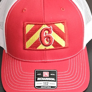 May include: Red and white trucker hat with a gold and red patch featuring the number "6". The hat has a white mesh back and a black brim. The hat has a Richardson label with the text "Est. 1870", "Richardson", "Original 112 Trucker", and "Adjustable".