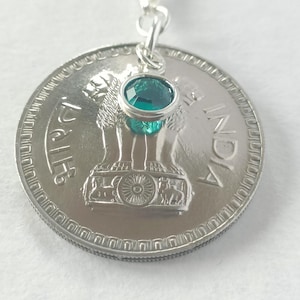 May include: Silver pendant necklace featuring a large coin with an Indian emblem. A small, oval-shaped, teal-coloured gemstone is suspended above the coin. The necklace chain is also silver.
