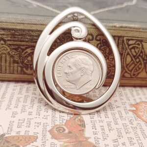 May include: Silver pendant necklace featuring a swirling design that holds a US dime coin. The coin is dated 2005 and has the inscription "In God We Trust". A small clear gemstone is set above the coin.