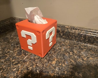 Op de afbeelding: Oranje tissuebox in de vorm van een vraagtekenblok uit het videospel Super Mario Bros. De doos heeft witte vraagtekens aan elke kant.