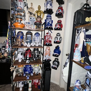 以下が含まれることがあります： 木製の棚に飾られたスターウォーズのR2-D2ロボットのコレクション。ロボットは赤、青、オレンジなど、さまざまな色をしています。一部のロボットは壁に掛けられています。