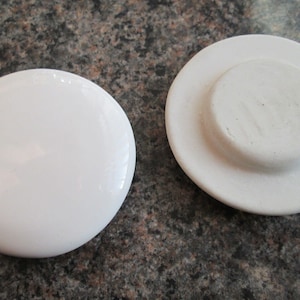 Puede incluir: Dos tapas de tarro de cerámica blanca, una plana y otra con un centro elevado. Las tapas son lisas y tienen un acabado ligeramente brillante.