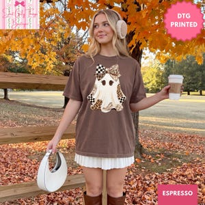 May include: A brown short-sleeve t-shirt featuring a graphic of a ghost wearing a large bow. The ghost design incorporates leopard print and checkered patterns. The shirt is paired with a white pleated skirt and a white crescent-shaped handbag.