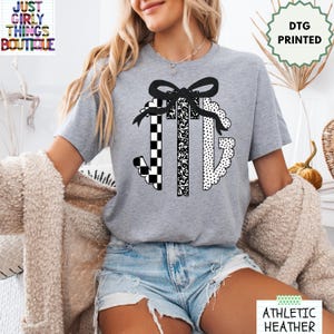 May include: A heather gray short-sleeve t-shirt featuring a black graphic print of a large monogram letter design topped with a bow. The monogram consists of three letters with distinct patterns: a checkerboard, a speckled texture, and polka dots.