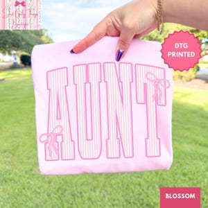 May include: A light pink t-shirt featuring the word AUNT in large, block-style letters with a white vertical pinstripe pattern. The letters are outlined in a darker pink, and two small pink ribbon bows are attached to the first and last letters.