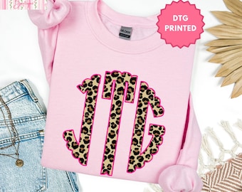 May include: A light pink crewneck sweatshirt featuring a large, central monogram with the letters JTG in a leopard print pattern, outlined in bright pink. The garment has a Gildan brand label at the inner neckline and a matching pink scrunchie.