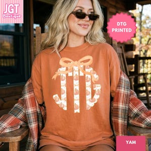 May include: A burnt orange short-sleeve t-shirt featuring a large central graphic of the letters JTG in a white font filled with a repeating pumpkin pattern, topped with a large peach-colored bow. The shirt is worn with a plaid flannel shirt.
