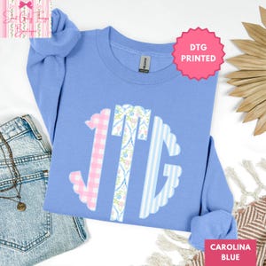 May include: Carolina blue sweatshirt with a DTG printed monogram. The monogram features a pink gingham letter, a floral letter, and a blue striped letter. The sweatshirt is next to a pair of blue jeans.