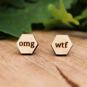 Könnte beinhalten: Ein Paar sechseckige Ohrringe aus Holz. Auf dem einen Ohrring steht "omg", auf dem anderen "wtf". Die Ohrringe sind hellbraun und liegen auf einer Holzoberfläche, mit grünen Blättern im Hintergrund.