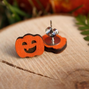 Puede incluir: Un par de pendientes de madera en forma de calabaza naranja con una cara sonriente tallada. Los pendientes tienen postes de plata.