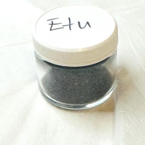 May include: A small clear glass jar with a white lid. The lid has the word "Etu" written on it in black marker. The jar is filled with a dark gray substance.