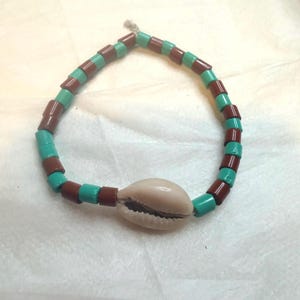 May include: A beaded bracelet featuring alternating cylindrical brown and turquoise beads strung together. A single, smooth, off-white cowrie shell is positioned as a central focal point on the strand, which is tied with a small knot at the top.
