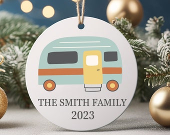 Op de afbeelding: Een witte keramische kerstversiering met een illustratie van een turquoise camper, met een oranje streep en een gele deur. De tekst "THE SMITH FAMILY 2023" staat eronder. Bovenop zit een touwlus.
