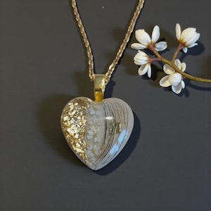 Könnte beinhalten: Eine herzförmige Anhängerkette an einer goldfarbenen Kette. Der Anhänger hat ein klares Harz-Finish mit Blattgoldflocken, kleinen weißen Blumen und feinen Haarsträhnen sowie einem eingebetteten goldfarbenen Buchstaben M auf der rechten Seite.