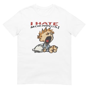 May include: White t-shirt featuring the text "I HATE MORNINGS!" in red and black. The graphic depicts a cartoon character yawning, wearing blue and white striped pyjamas, and sitting on a pillow.