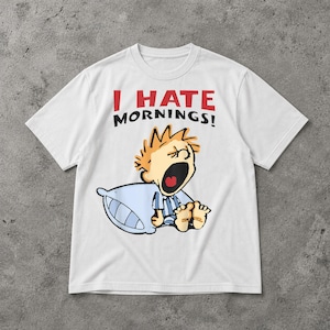 May include: White t-shirt featuring the text "I HATE MORNINGS!" in red and black. The graphic depicts a cartoon character with orange hair, yawning, holding a blue pillow. The character is wearing blue pyjamas.