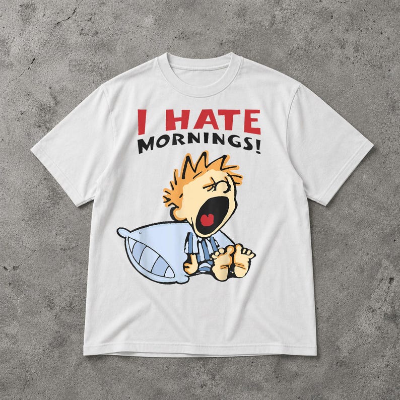 May include: White t-shirt featuring the text "I HATE MORNINGS!" in red and black. The graphic depicts a cartoon character with orange hair, yawning, holding a blue pillow. The character is wearing blue pyjamas.