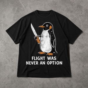 Könnte beinhalten: Schwarzes T-Shirt mit einem Cartoon-Pinguin, der ein Messer hält. Der Pinguin hat einen weißen Bauch, einen schwarzen Rücken und orangefarbene Füße und einen Schnabel. Der Text "FLIGHT WAS NEVER AN OPTION" ist darunter gedruckt.