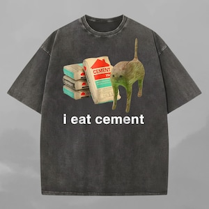 Op de afbeelding: Donkergrijze t-shirt met een afbeelding van een kat en cementzakken. De tekst "i eat cement" is in het wit gedrukt. Het shirt heeft een ronde hals en korte mouwen.