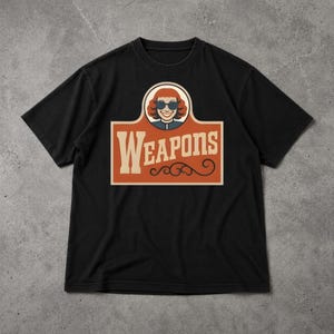 Op de afbeelding: Zwart t-shirt met een retro graphic. Het ontwerp toont een lachende vrouw met een zonnebril, boven het woord "WEAPONS" in een vintage lettertype, op een rechthoekige achtergrond.