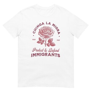 Op de afbeelding: Wit t-shirt met een bordeauxrode roos en de tekst "CHINGA LA MIGRA" erboven. Daaronder staat "Protect & Defend IMMIGRANTS". Het t-shirt heeft een ronde hals en korte mouwen.