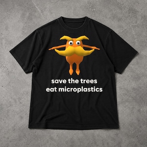 Op de afbeelding: Een zwart T-shirt met korte mouwen en een afbeelding van een oranje, cartoonachtig wezen met een grote gele snor en grote ogen. Onder het wezen staat de witte tekst: save the trees eat microplastics in een eenvoudig lettertype.