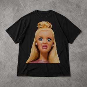 Könnte beinhalten: Schwarzes T-Shirt mit dem Gesicht einer Puppe mit überraschtem Ausdruck. Die Puppe hat blondes Haar, blaue Augen und trägt ein rosa Oberteil. Das T-Shirt ist aus einem weichen Material.
