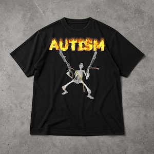 Puede incluir: Camiseta negra con la palabra "AUTISMO" en una fuente llameante sobre una figura de esqueleto que sostiene dos pistolas. La camiseta gráfica tiene cuello redondo y mangas cortas. La camiseta está hecha de un material suave.