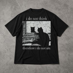 Op de afbeelding: Zwart t-shirt met een witte afbeelding van twee katten, waarvan één in een spiegel, en de tekst "i do not think therefore i do not am".