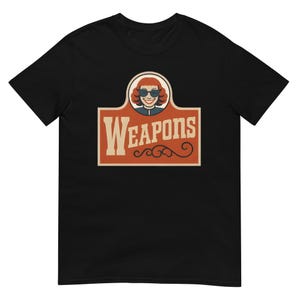May include: Black t-shirt featuring a retro-style graphic. The graphic includes a woman wearing sunglasses and the word "WEAPONS" in a vintage font. The design is in shades of red, beige, and brown.