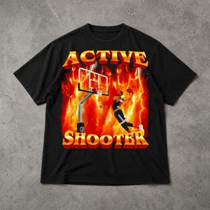 Może przedstawiać: Czarna koszulka z grafiką o tematyce koszykówki. Projekt przedstawia koszykarza wsadzającego piłkę do kosza, kosz i słowa "ACTIVE SHOOTER" w płomiennej, retro czcionce. Tło ma płomienie i błyskawice.