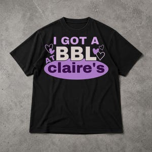 Op de afbeelding: Zwart T-shirt met de tekst "I GOT A BBL at claire's" in wit en paars. Het ontwerp bevat paarse harten en een ovaal. Het T-shirt heeft een ronde hals en korte mouwen.