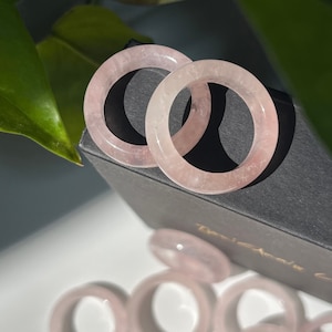 Op de afbeelding: Een set roze rozenkwarts ringen, gestapeld op elkaar, in een zwarte doos. De ringen zijn glad en gepolijst, met een subtiele glans.