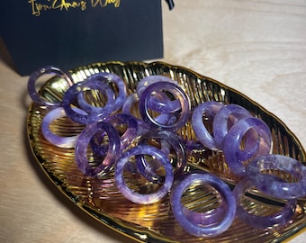 May include: A gold-colored dish filled with amethyst rings. The rings are a light purple color and have a smooth, polished finish.