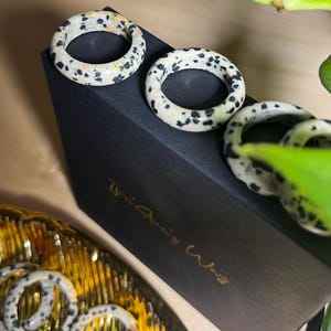 May include: Four black and white speckled rings on a black box with the text "The Artist Way" in gold lettering. The rings are made of natural stone and have a unique, organic look.