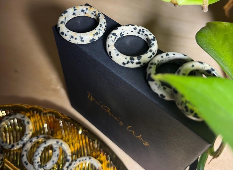 May include: Four black and white speckled rings on a black box with the text "The Artist Way" in gold lettering. The rings are made of natural stone and have a unique, organic look.