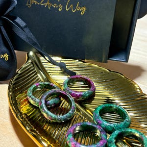 May include: Five circular rings crafted from a speckled green and magenta stone material, arranged on a gold-colored leaf-shaped tray. The rings feature a polished finish with visible mineral inclusions, resting near a black box with gold text.