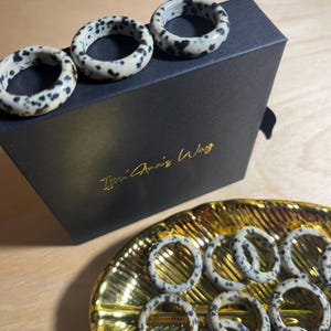 May include: A set of dalmatian jasper rings in various sizes, displayed on a black box with the text "Tsoni Anais Way" and a gold tray. The rings are made of natural stone with a white and black speckled pattern.