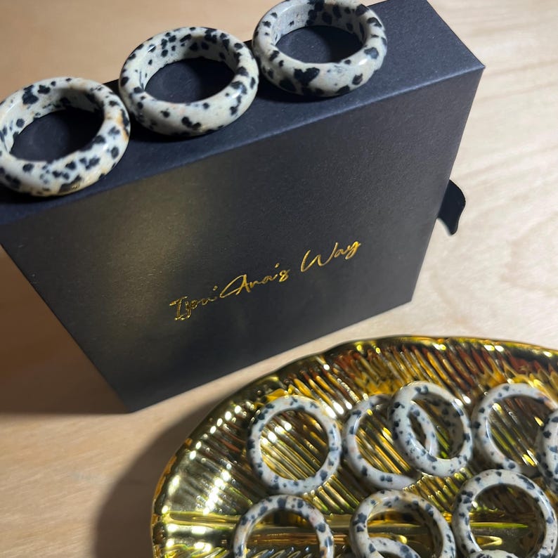 May include: A set of dalmatian jasper rings in various sizes, displayed on a black box with the text "Tsoni Anais Way" and a gold tray. The rings are made of natural stone with a white and black speckled pattern.