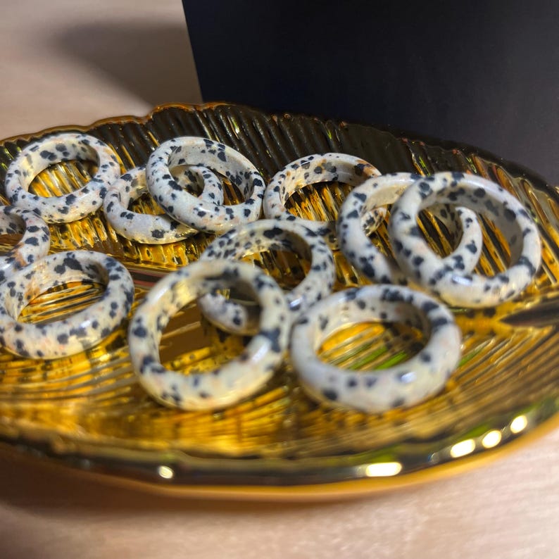 May include: A collection of dalmatian jasper rings in various sizes, displayed on a gold leaf-shaped dish. The rings are made of natural stone with a white base and black spots.