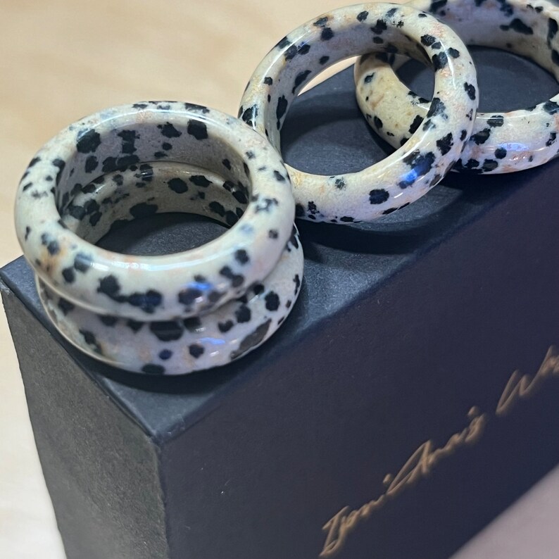 May include: Four stacked rings made of dalmatian jasper, a white stone with black spots. The rings are on a black box with the text "Zenith Arts" in gold lettering.