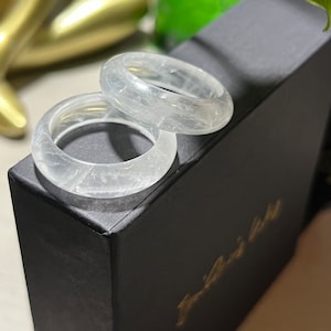 Peut inclure: Deux bagues en cristal blanc transparent avec une apparence nuageuse et texturée. Les bagues sont empilées l'une sur l'autre et reposent sur une boîte noire.