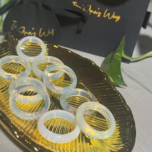 Könnte beinhalten: Eine Sammlung von acht regenbogenfarbenen Opalringen auf einer goldenen Blattoberfläche. Die Ringe sind übereinander gestapelt und in einem Kreis angeordnet.