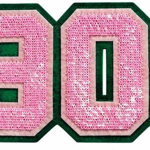May include: A large iron-on patch featuring the numbers 1908 in a block font. The numbers are covered in light pink sequins with a light pink felt border, all mounted on a dark green felt backing that follows the shape of the digits.