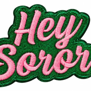 May include: An embroidered patch featuring the text Hey Soror in pink cursive lettering. The letters are set against a dark green background with a scalloped edge, creating a distinct contrast between the pink thread and the green fabric base.