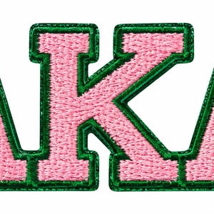 May include: An embroidered patch featuring the letters AKA in a bold, block-style font. The letters are filled with light pink thread and are outlined with a thick, dark green border, creating a high-contrast, textured appearance.