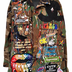 May include: A camouflage print button-down shirt featuring colorful paint splatters and various embroidered patches, including a large black logo, a rainbow peace sign, a rainbow lip design, and text reading "CUTE BUT PSYCHO" and "LIKE MY LIQUOR STRAIGHT."