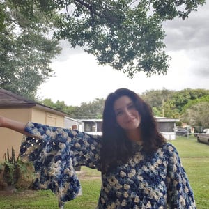 May include: A handmade crochet top in shades of blue and cream. The top features a square pattern and flared sleeves. The person in the image is wearing the top outdoors in front of a green lawn and trees.