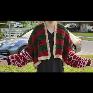 May include: Crocheted cardigan in red and green stripes with beige trim. The sleeves feature a unique pattern in red and pink. The cardigan has distressed details, adding a unique touch to this handmade garment.