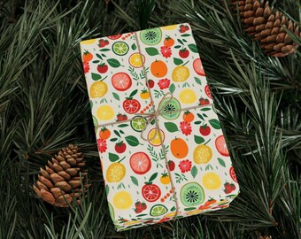 May include: A gift box wrapped in white paper with a colorful fruit pattern. The paper features lemons, limes, oranges, strawberries, kiwi, and red and orange flowers. The box is tied with twine.